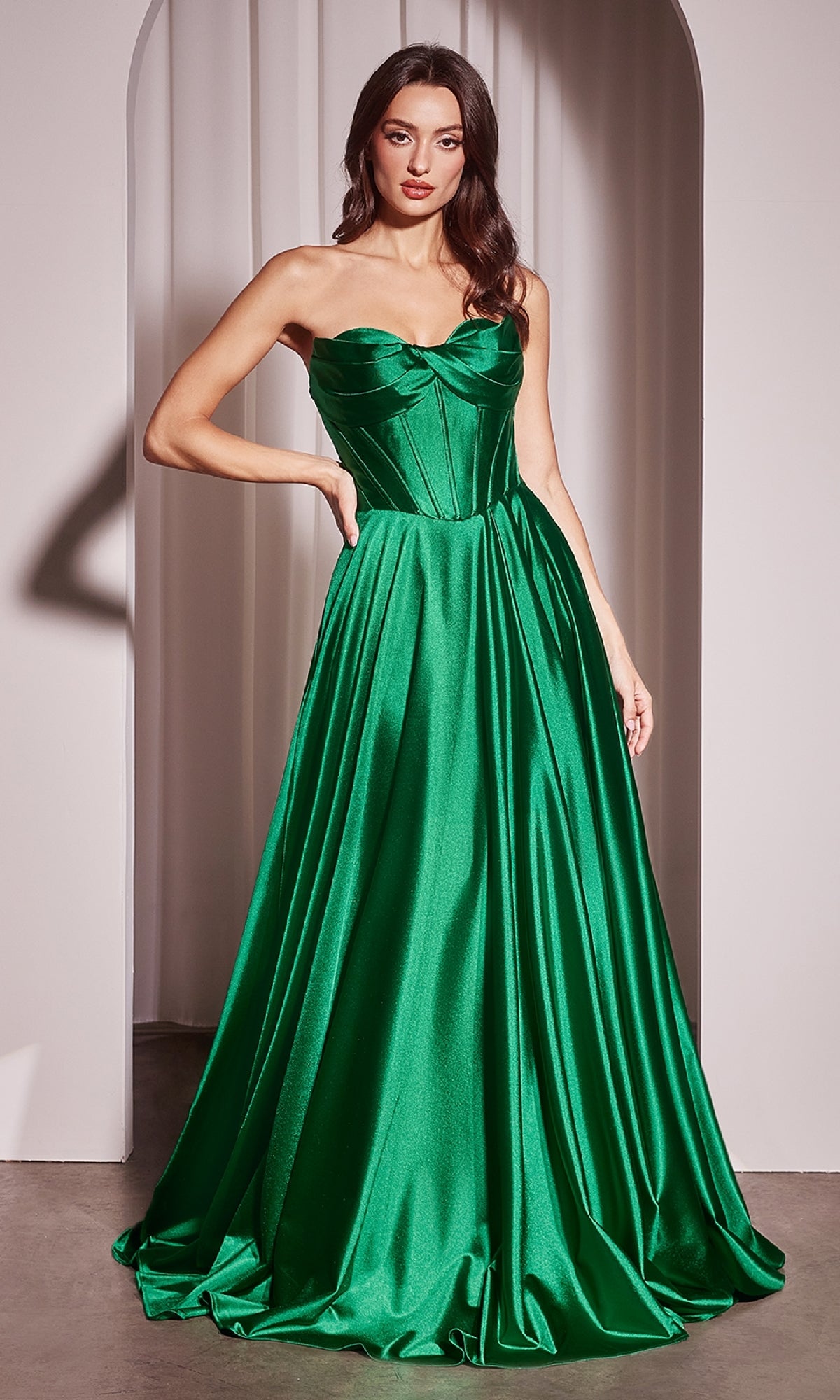 A woman stands in an arched doorway wearing the Ladivine CD378, a strapless long A-line prom dress in emerald satin with a corset-style bodice and gracefully flared skirt.